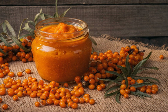 Sea buckthorn puree in a glass jar surrounded by fresh orange sea buckthorn berries on a rustic burlap background.